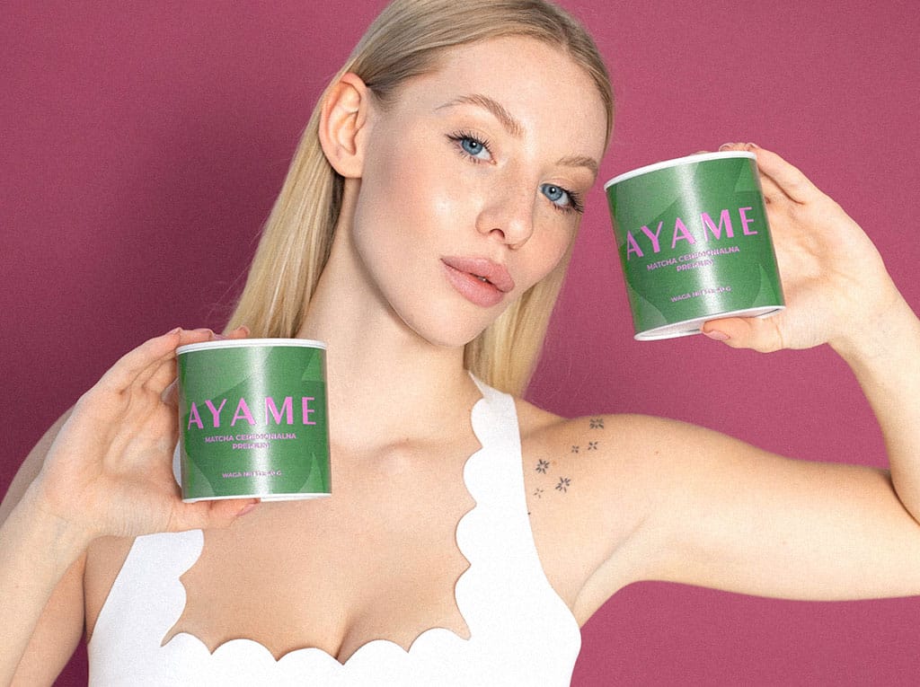 Blondynka prezentująca dwie puszki Matcha Ceremonialna Premium Ayame na różowym tle – japońska herbata z Uji.
