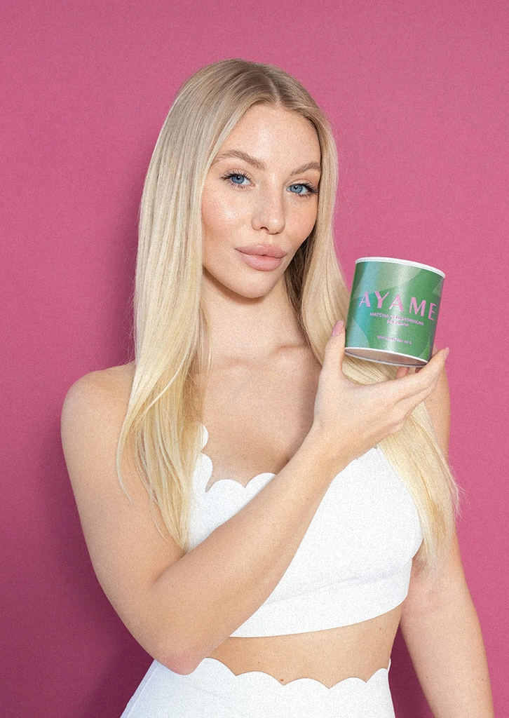 Blondynka w białym topie trzyma puszkę Matcha Ceremonialna Premium Ayame Matcha na różowym tle.